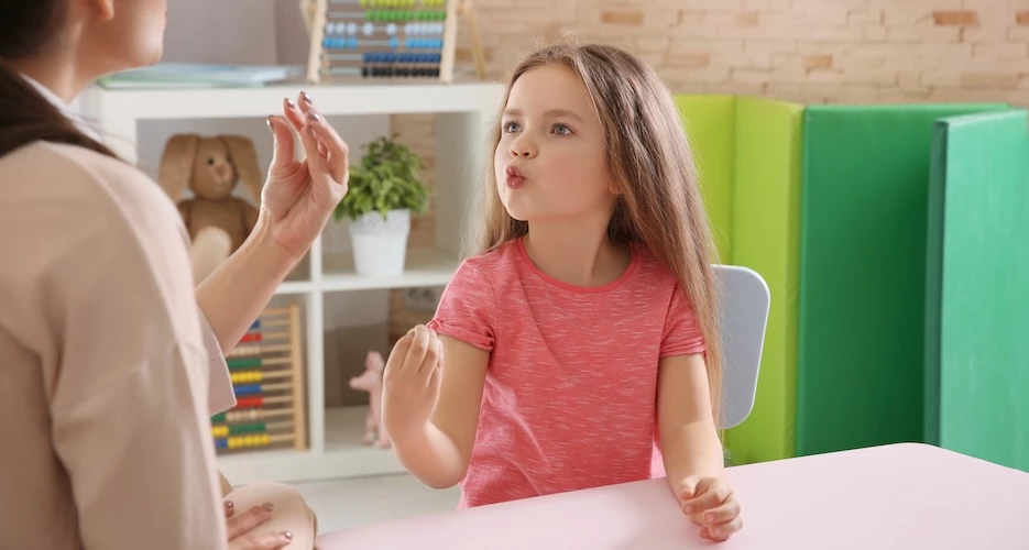 terapeuta da fala faz exercícios com uma menina de aproximadamente 7 anos, vestida com t-shirt rosa. 